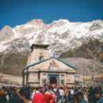 A crowd of pilgrims at Kedarnath Temple with snowcapped Himalayan peaks in the background.