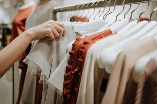 A person browsing neutral and terracotta clothing hanging on white hangers in a men's and women's cloth shop in khatima